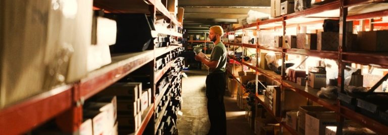 Pessoa em um depósito ou almoxarifado iluminado, cercada por prateleiras metálicas cheias de caixas e pacotes. A pessoa, usando uma touca amarela e roupas casuais, consulta um dispositivo ou uma prancheta enquanto organiza ou inspeciona os itens armazenados