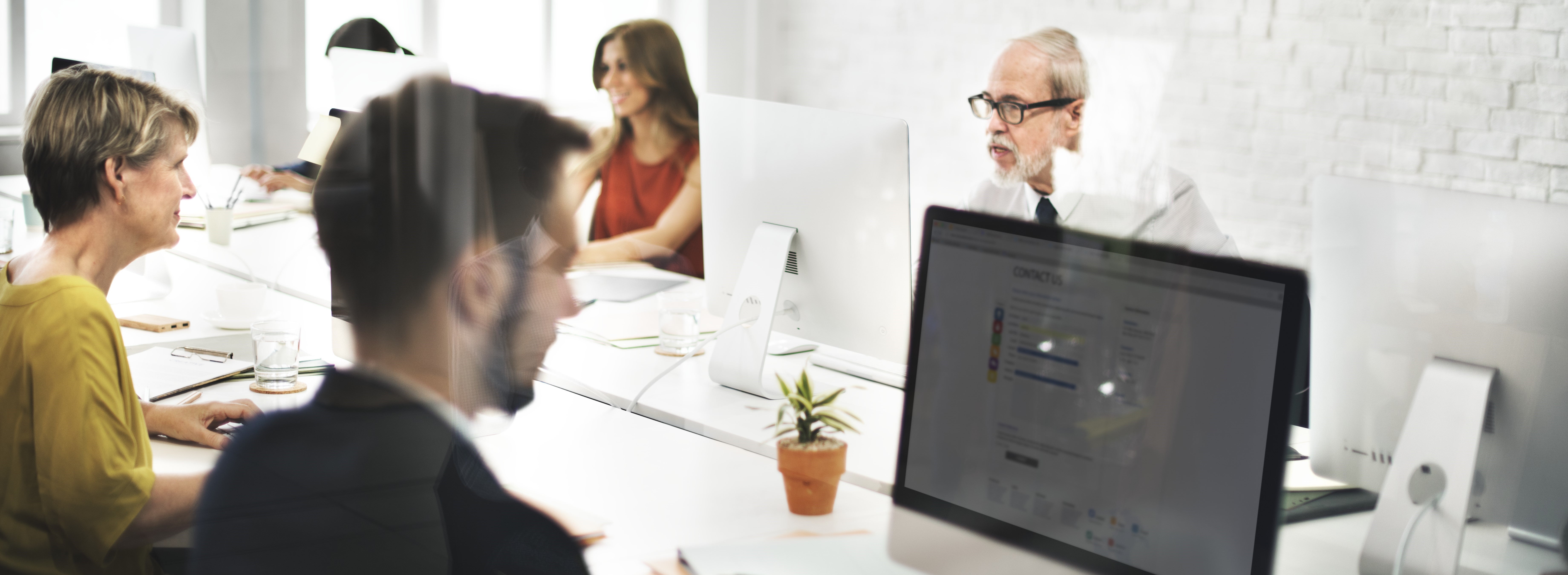 Equipe de profissionais trabalhando em um escritório moderno com computadores, interagindo de forma colaborativa em um ambiente claro e bem iluminado.