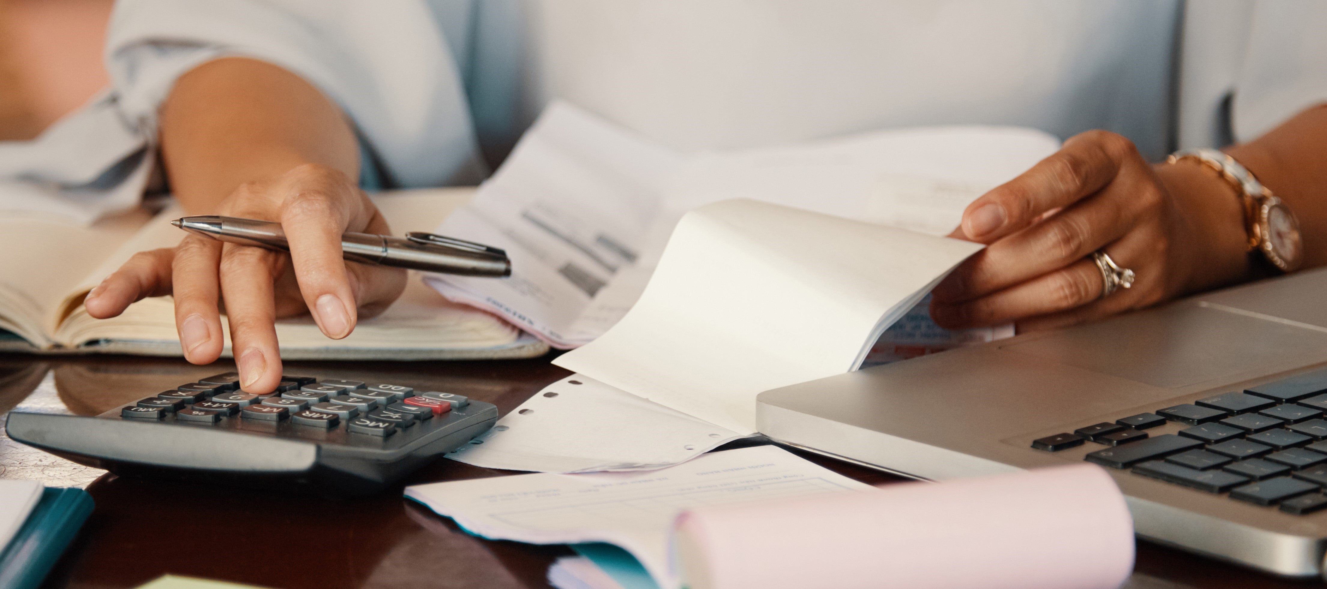 Pessoa sentada à mesa trabalhando com calculadora, laptop, papéis e cadernos. A pessoa segura uma caneta e manuseia papéis, aparentemente revisando contas ou relatórios financeiros. Há também notas adesivas coloridas na mesa