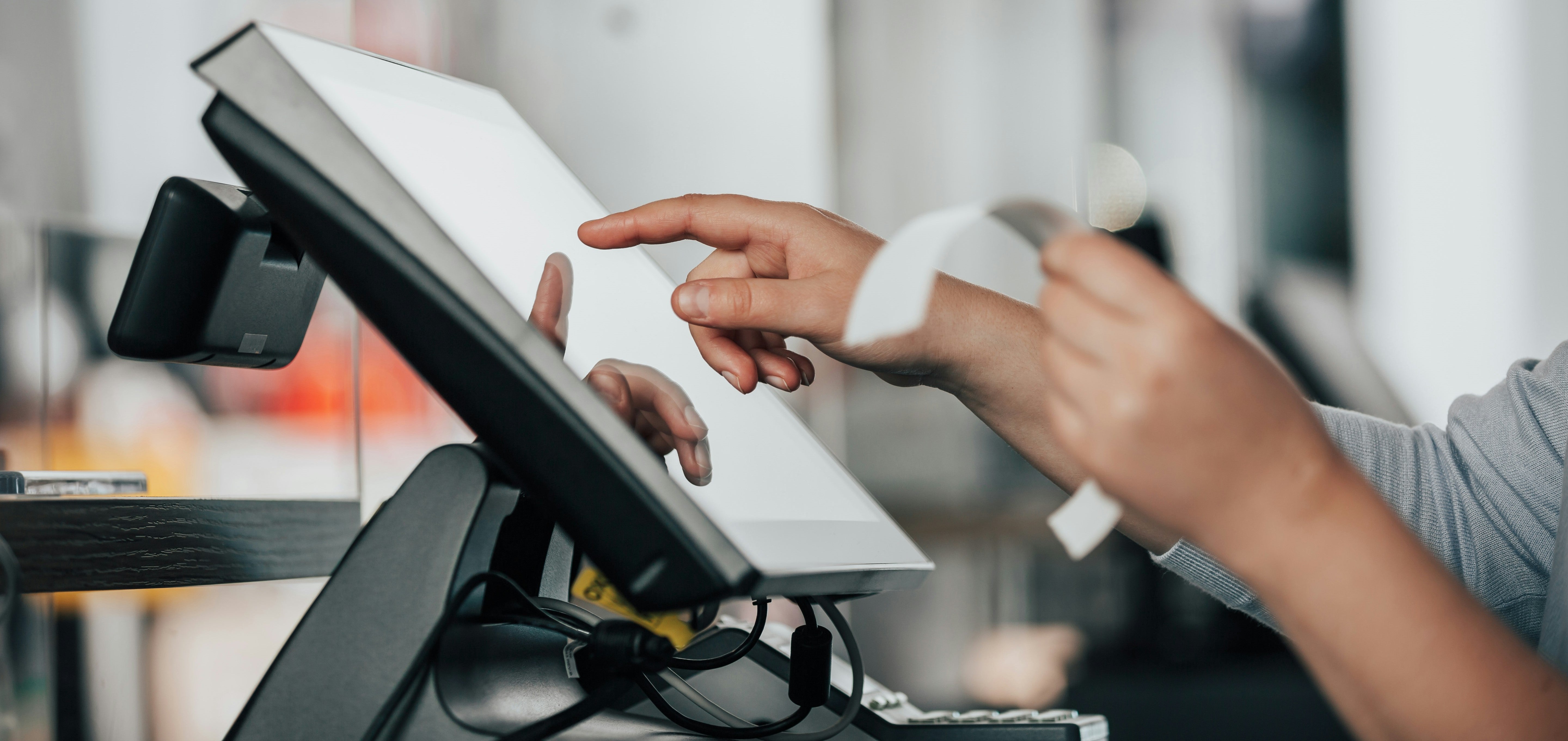 Pessoa utilizando uma tela sensível ao toque em um terminal de ponto de venda (PDV), enquanto segura um recibo impresso na outra mão. A cena mostra o ambiente típico de um caixa de loja ou restaurante