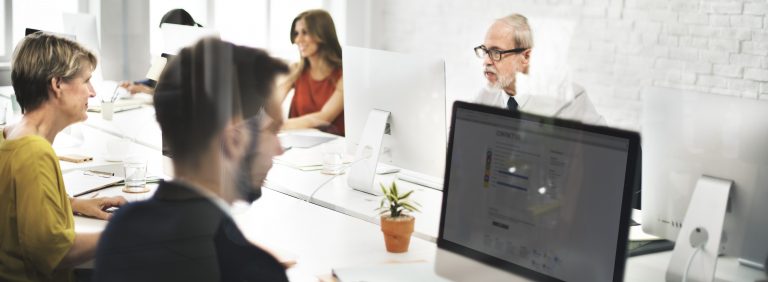 Equipe de profissionais trabalhando em um escritório moderno com computadores, interagindo de forma colaborativa em um ambiente claro e bem iluminado.