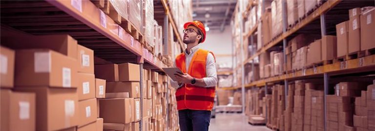 Profissional com colete e capacete no centro de distribuição, usando sistema de gestão para organizar estoque e pedidos.